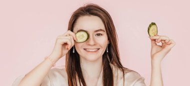 Kız, ev yapımı güzellik maskeleri yapıyor. Salatalık gözün etrafındaki derinin tazeliği için. Kadın genç cildine dikkat eder. Model gülüyor ve pembe arka planda kaplıcada eğleniyor.