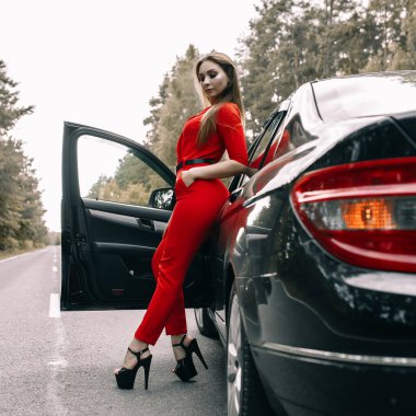 Kırmızı tulumlu güzel bir genç kız ormanda boş bir yolda siyah bir arabanın yanında duruyor.
