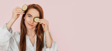 Kız, ev yapımı güzellik maskeleri yapıyor. Salatalık gözün etrafındaki derinin tazeliği için. Kadın genç cildine dikkat eder. Model gülüyor ve pembe arka planda kaplıcada eğleniyor.