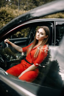 Kırmızı tulumlu güzel bir genç kız ormanda boş bir yolda siyah bir arabanın direksiyonunda oturuyor.