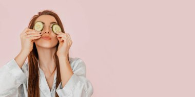 Kız, ev yapımı güzellik maskeleri yapıyor. Salatalık gözün etrafındaki derinin tazeliği için. Kadın genç cildine dikkat eder. Model gülüyor ve pembe arka planda kaplıcada eğleniyor.