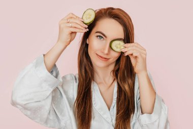 Kız, ev yapımı güzellik maskeleri yapıyor. Salatalık gözün etrafındaki derinin tazeliği için. Kadın genç cildine dikkat eder. Model gülüyor ve pembe arka planda kaplıcada eğleniyor.