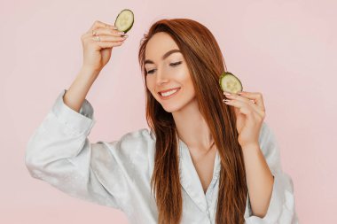 Kız, ev yapımı güzellik maskeleri yapıyor. Salatalık gözün etrafındaki derinin tazeliği için. Kadın genç cildine dikkat eder. Model gülüyor ve pembe arka planda kaplıcada eğleniyor.