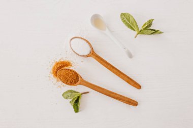 Tahta kaşıklar beyaz, kamış şekeri ve beyaz mutfak masasında nane yaprakları. Tatlandırıcılar yemek malzemeleri