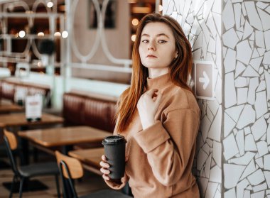 Tarz sahibi kız bir kafede kahve içiyor. Kızıl saçlı bir kadın bej takım elbiseli bir duvarın önünde duruyor. Modern iç mekan. Kahve karton bardağa konacak.