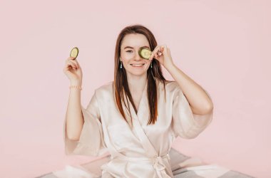 Kız, ev yapımı güzellik maskeleri yapıyor. Salatalık gözün etrafındaki derinin tazeliği için. Kadın genç cildine dikkat eder. Model gülüyor ve pembe arka planda kaplıcada eğleniyor.