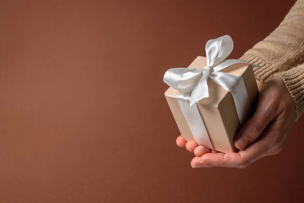 Beige gift box with white bow in woman hands on brown background