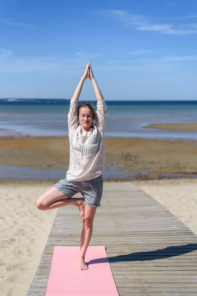 Woman in linen clothes doing yoga tree pose on beach at sunny day. Wellbeing, physical and mental health