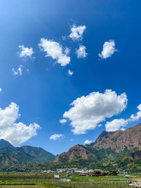 Bir vadi ve tepelerin üzerinde yeşil bitkilerle kaplı beyaz bulut kümeleri olan açık mavi bir gökyüzü..