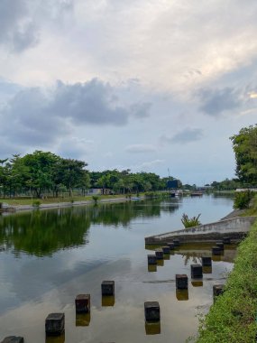 Suyun üzerinde ağaç yansımaları olan sakin bir kanal manzarası, küp şeklinde beton sütunlar ve suyun kenarında yüzen bir duba..