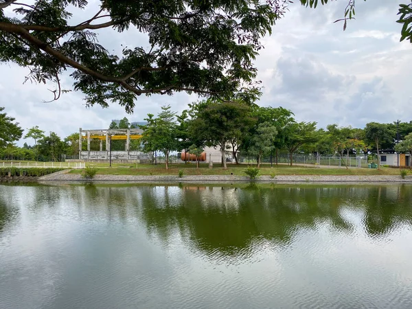 View of a canal bank with tree reflections, featuring a yellow gantry or industrial facility structure in the distance.