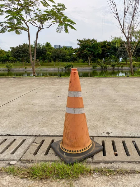 Dirty, old orange traffic cone with two white reflective stripes, placed over a concrete drain cover on the edge of a pathway.