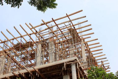 İnşaat halindeki çok katlı bir binanın üst katındaki beton çerçevenin etrafındaki ahşap iskeleler ve çelik destek kuvvetleri mavi gökyüzüne karşı..