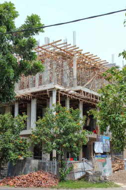İnşaat halindeki çok katlı binanın dış görünüşü, ahşap iskelelerle çevrili, ön planda gür ağaçlar var..