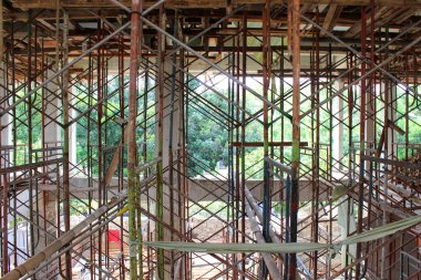 Bir inşaat sahasında çok renkli metal boru ve ahşap iskelelerden oluşan yoğun bir ağ, pencere çerçevesi dışında yemyeşil ağaçlardan oluşan bir arka plan veya açık.