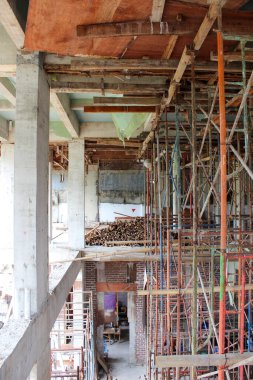 İnşaat halindeki bir binanın güçlendirilmiş beton kolonu ve kiriş yapısı, paslı metal boru iskelesi üst kattaki döküm işini destekliyor..