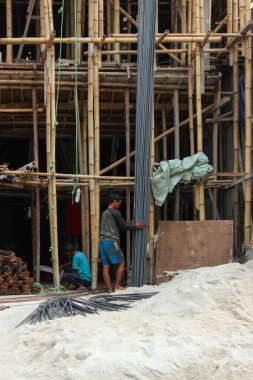 Jakarta, Endonezya - 11 Aralık 2025: Bambu iskelesinin yanında uzun demir çubuklar ve bir ev inşaatında kum yığını taşıyan iki inşaat işçisi.