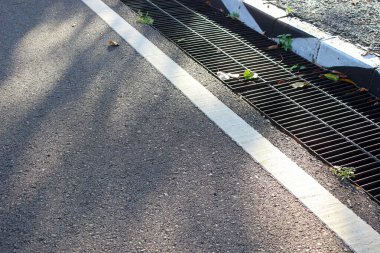 Beyaz bir çizginin yanında kaba asfalt dokusu ve halka açık bir alanda güneş ışığıyla aydınlatılmış siyah bir metal ızgarayla kaplanmış bir drenaj çukuru..