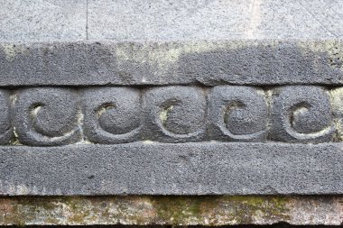 Bir heykel tabanında, sarımsı yeşil yosunlu ya da liken taşı yüzeyinde tekrarlayan bir su girdabı ya da ceplok deseni olan taş kalıntısı var..