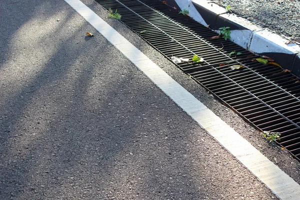 Beyaz bir çizginin yanında kaba asfalt dokusu ve halka açık bir alanda güneş ışığıyla aydınlatılmış siyah bir metal ızgarayla kaplanmış bir drenaj çukuru..