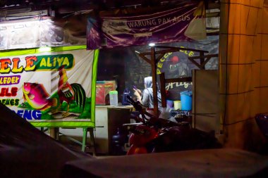 Cakarta, Endonezya - 20 Aralık 2025: Bir kadın Pecel Lele Aliya tezgahında oturuyor ve önünde parlak bir bayrak var.