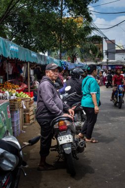 Jakarta, Endonezya - 20 Aralık 2025: Gri kapüşonlu orta yaşlı bir adam geleneksel pazar kalabalığının ortasında Honda motosikletinin üzerinde oturuyor.