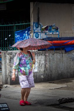Cakarta, Endonezya - 20 Aralık 2025: Renkli kıyafetler giyen yaşlı bir kadın güneşin sıcağından korunmak için ekose bir şemsiye kullanarak yürüyor.