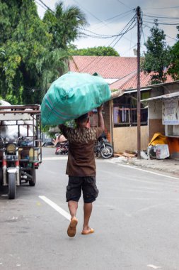 Cakarta, Endonezya - 20 Aralık 2025: Bir adam yerleşim yerindeki bir sokakta kafasında atık veya plastik çöp dolu büyük bir çuval taşır.