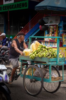 Jakarta, Endonezya - 20 Aralık 2025: Cengkareng pazar bölgesinde muz ve papayaları satmak için bisiklet arabası kullanan yaşlı bir meyve satıcısı.