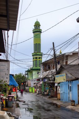 Jakarta, Endonezya - 18 Ocak 2026: Bojong Raya 'da yeşil cami minaresi ve dağınık elektrik hatları olan dar bir sokak manzarası.