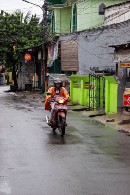 Jakarta, Endonezya - 18 Ocak 2026: Turuncu yağmurluk giyen bir motosikletli yeşil bir binanın önünde ıslak bir yoldan geçiyor.