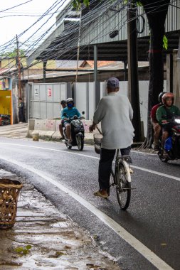 Jakarta, Endonezya - 18 Ocak 2026: Bulutlu hava şartları sırasında yol kenarında eski bir bisiklete binen yaşlı bir adam elektrik hatlarını geçiyor.