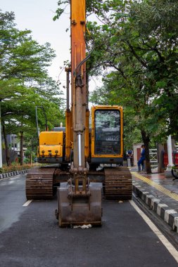 Jakarta, Endonezya - 18 Ocak 2026: Yeşil ağaçlı bir yaya bölgesinde duran sarı kazıcı makinesinin ön görüntüsü.