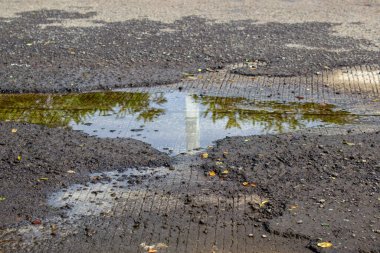 Asfalt yolundaki çukur, inşaat alanındaki ağaçları yansıtan yağmur suyuyla dolu..