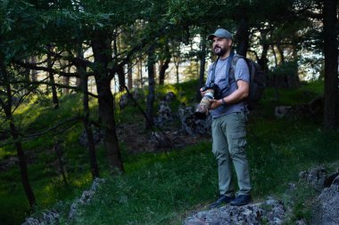 Ormandaki doğa fotoğrafçısı telefoto lensi ve sırtında bir kamera çantasıyla. Adam etrafındaki hayvanları fotoğraflamak için doğayı inceliyor..