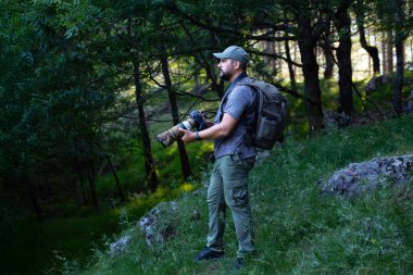 Ormandaki doğa fotoğrafçısı telefoto lensi ve sırtında bir kamera çantasıyla. Adam etrafındaki hayvanları fotoğraflamak için doğayı inceliyor..
