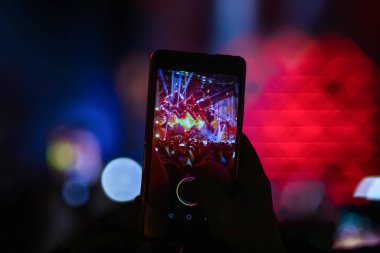 Geceleri canlı müzik performansı kaydetmek için cep telefonu tutan bir elin yakın plan çekimi. Telefon ekranı sahneyi parlak ışıklar ve kalabalıkla gösterirken, arka planda güzel kırmızı ve mavi Bokeh ışık efektleri yer alıyor..