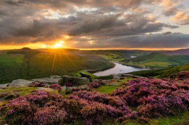 Akşam güneşi dramatik bulutların arasından yükseliyor. Tepe bölgesindeki Ladybower Barajı destansı manzarasıyla..