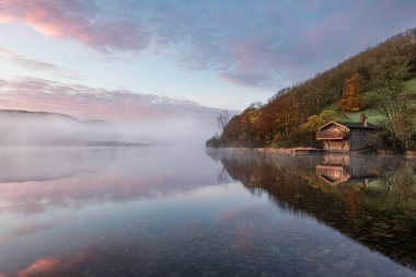 Ullswater Gölü 'ndeki kayıkhanede şafak vakti gökyüzünde güzel bir güneş doğumu rengi, göl sisi ve sakin yansımaları..