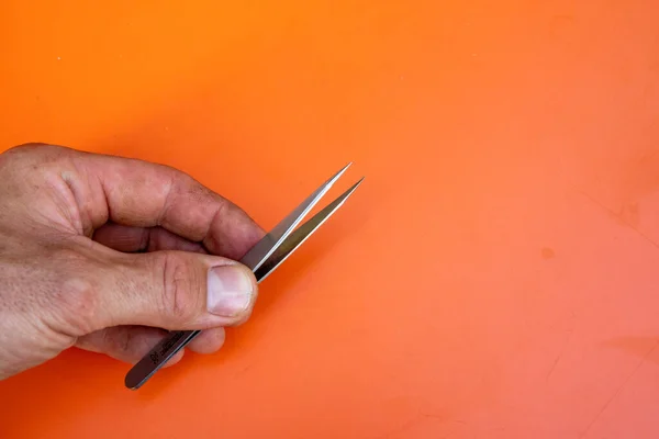 Male hand with tweezers in front of orange background. Copy Space on the right side. — Stock Photo, Image
