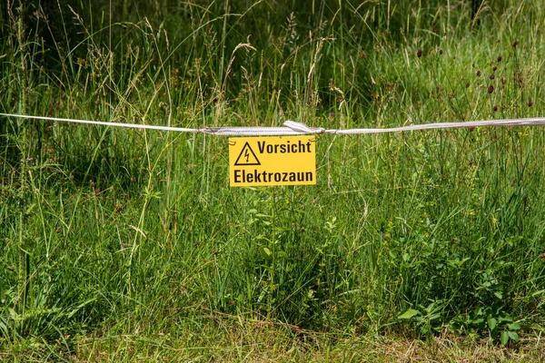 Εγγραφείτε με την επιγραφή στη γερμανική προσοχή Ηλεκτρικός φράχτης κρέμεται σε ένα ηλεκτρικό φράχτη μπροστά από ένα πράσινο λιβάδι — Φωτογραφία Αρχείου