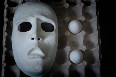 A white scary theater mask lies next to chicken eggs on a dark background clipart