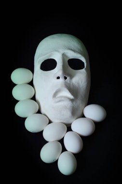 Beyaz, korkunç bir tiyatro maskesi, arka planda tavuk yumurtalarının yanında duruyor.