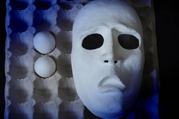 A white scary theater mask lies next to chicken eggs on a dark background