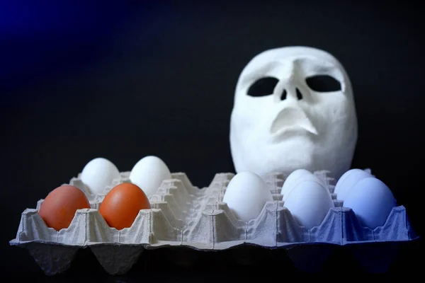 A white scary theater mask lies next to chicken eggs on a dark background
