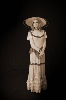 Eski elbiseli ve şapkalı bir kız koyu bir arka planda duruyor ve üzgün.