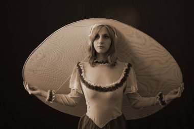 Eski elbiseli ve şapkalı bir kız koyu bir arka planda duruyor ve üzgün.