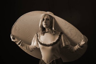 Eski elbiseli ve şapkalı bir kız koyu bir arka planda duruyor ve üzgün.