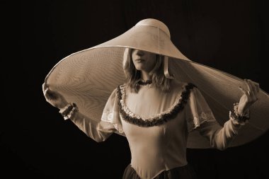 Eski elbiseli ve şapkalı bir kız koyu bir arka planda duruyor ve üzgün.