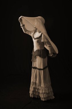 Eski elbiseli ve şapkalı bir kız koyu bir arka planda duruyor ve üzgün.